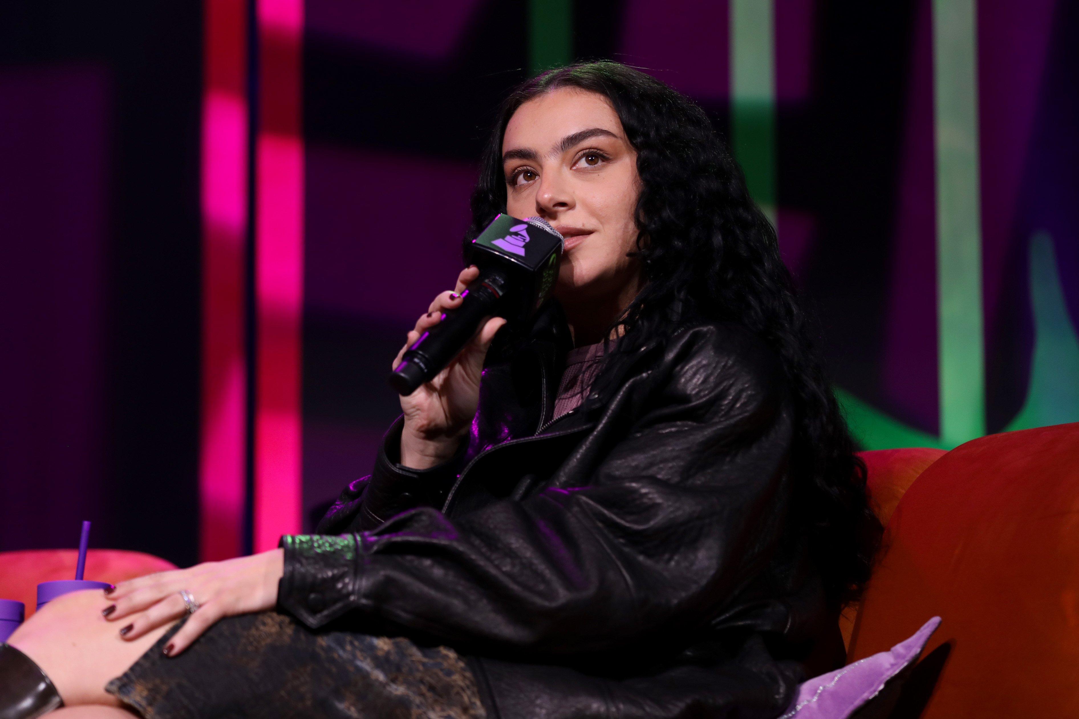 Photo of Charli XCX speaking at the 2024 GRAMMY U Fall Summit. Charli XCX is wearing a black leather jacket and holding a black microphone featuring the GRAMMY U logo